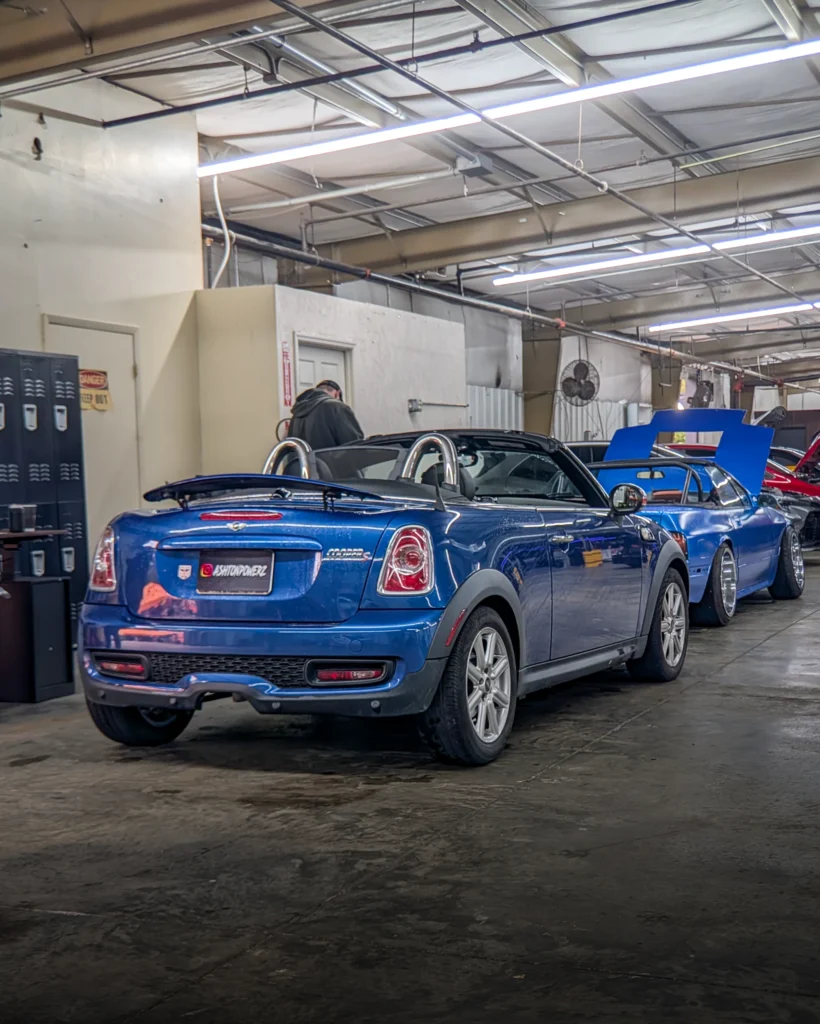 Blue Mini Cooper S convertible parked indoors with a blue coupe next to it and a person standing by a wall