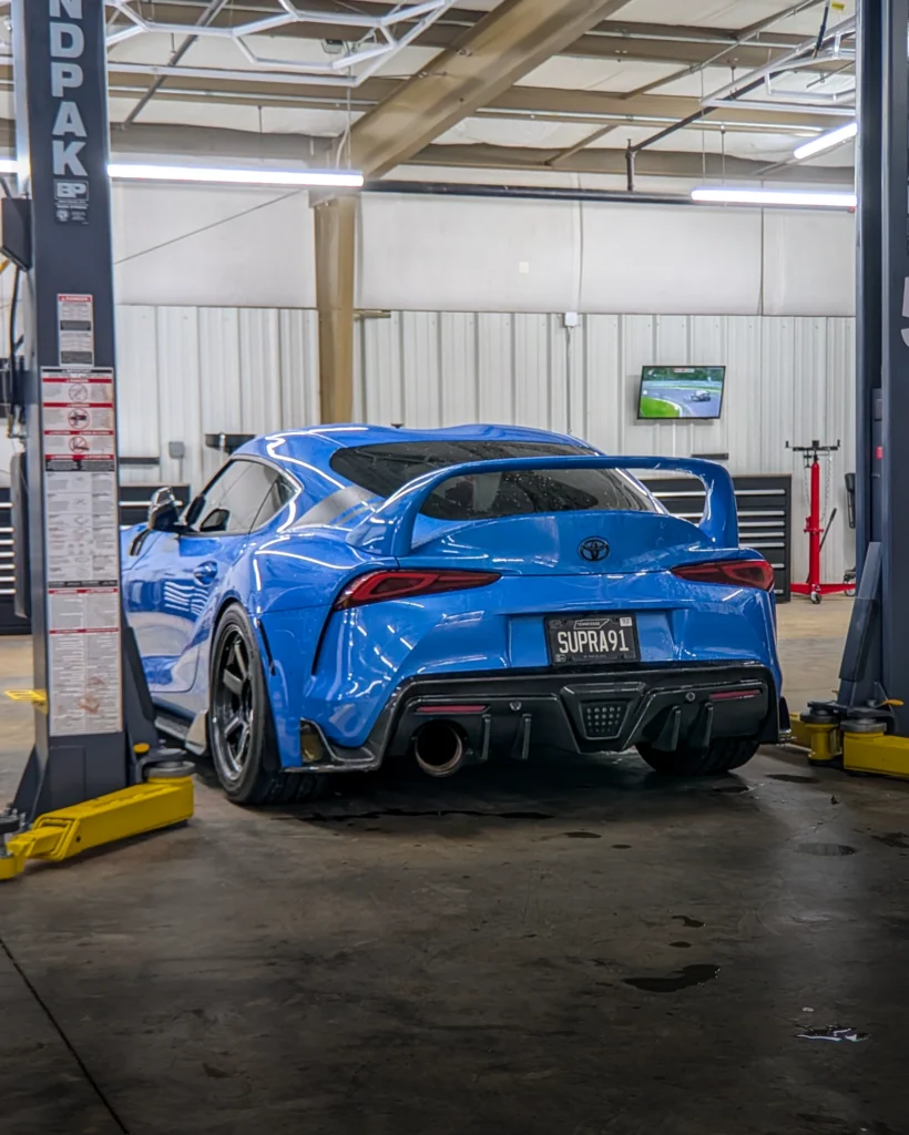 Blue Toyota Supra with spoiler in an indoor garage with car lift pillars and a TV screen in the background