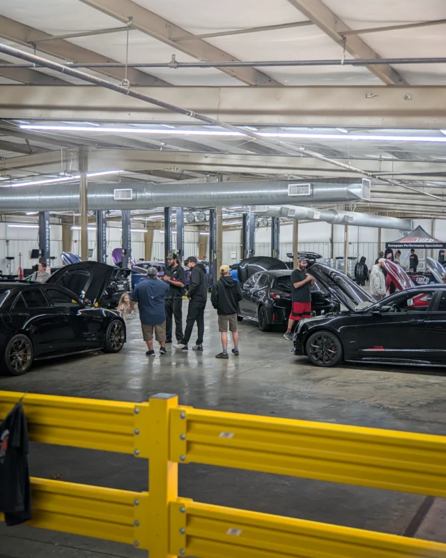 Group of people standing and talking near black cars inside a garage with yellow barriers and exposed ceiling ducts.