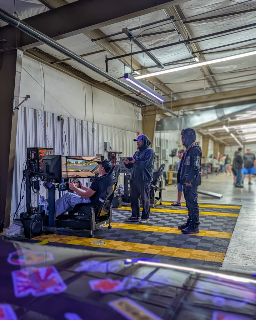 People playing a racing simulator indoors at an automotive event with a vehicle windshield covered in stickers in foreground.