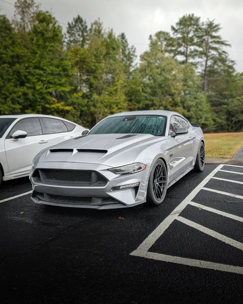 Silver Ford Mustang GT parked next to a white sedan on a wet day with trees in the background