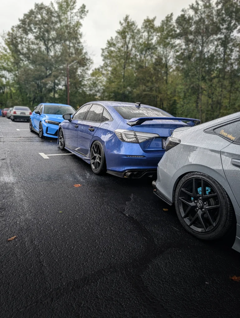 Three parked Honda sedans in gray, blue, and light blue on a wet asphalt surface with trees in the background