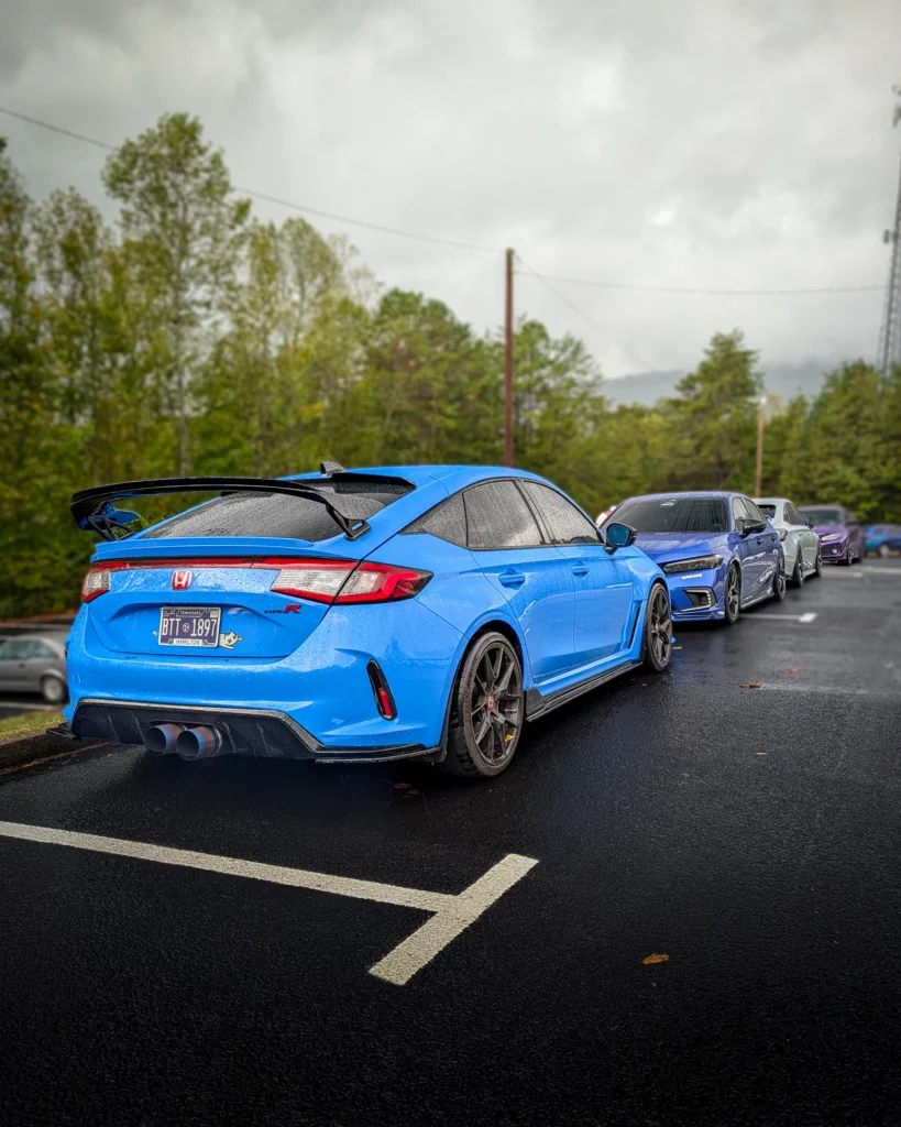 Blue Honda Civic Type R parked on wet asphalt with three more cars in a row on a cloudy day near trees