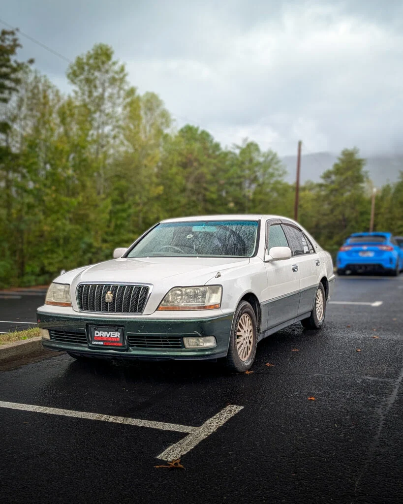 White Toyota Crown sedan parked on wet pavement in an outdoor setting with trees in background and a blue Porsche 911 in