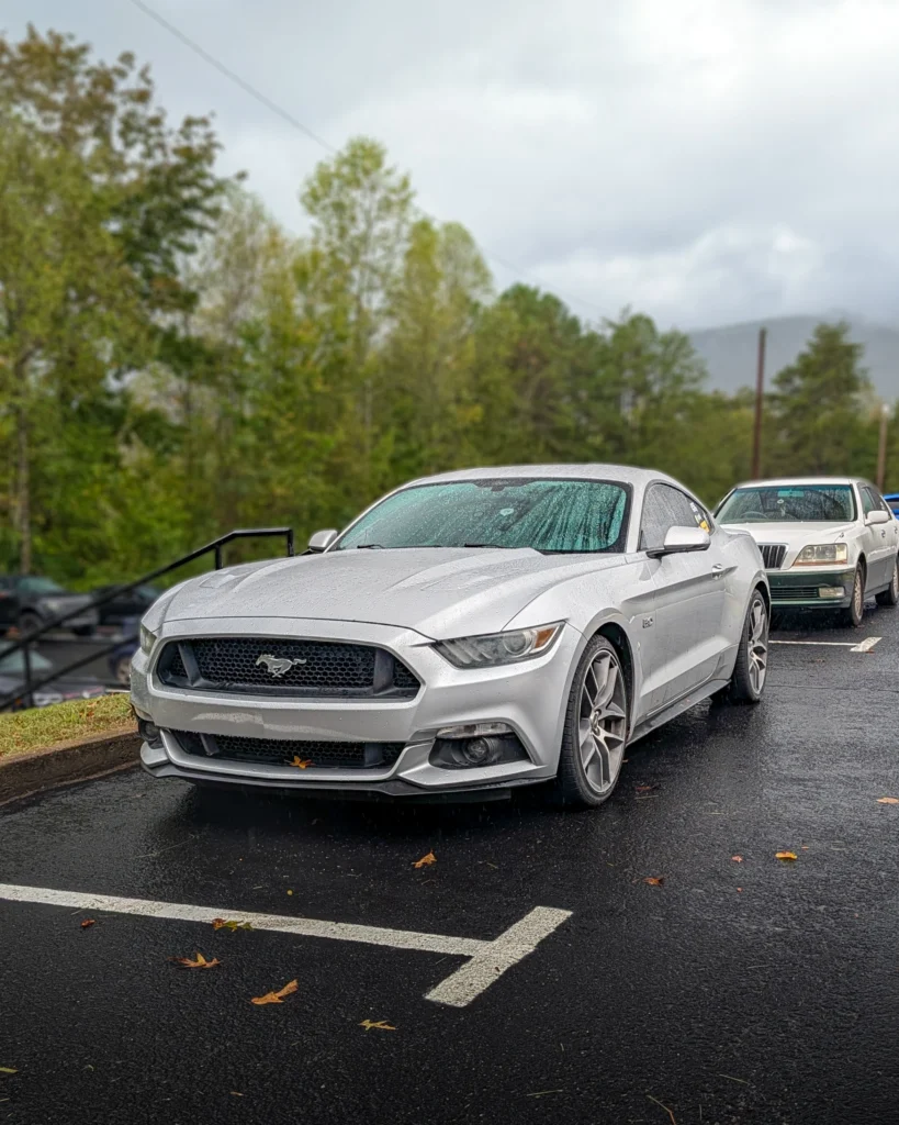 Silver Ford Mustang GT parked outdoors on wet asphalt with a silver sedan behind, surrounded by trees and an overcast sky.