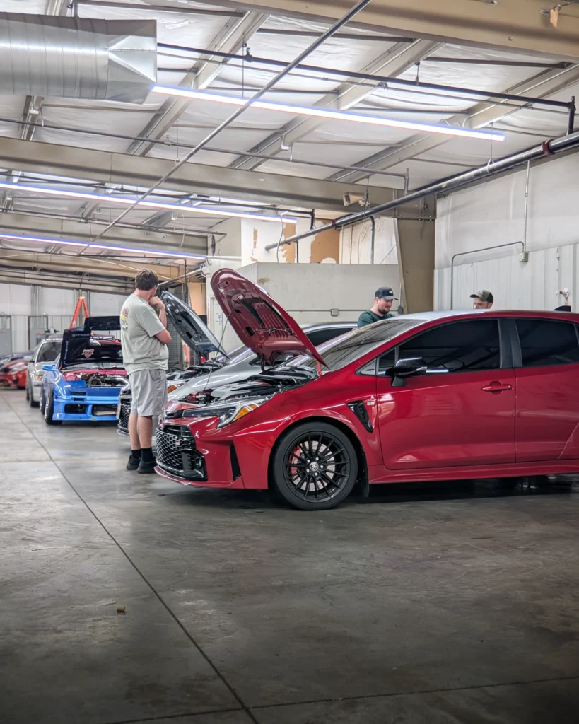 Red Toyota GR Corolla and two other cars with open hoods inside a garage with three men around them