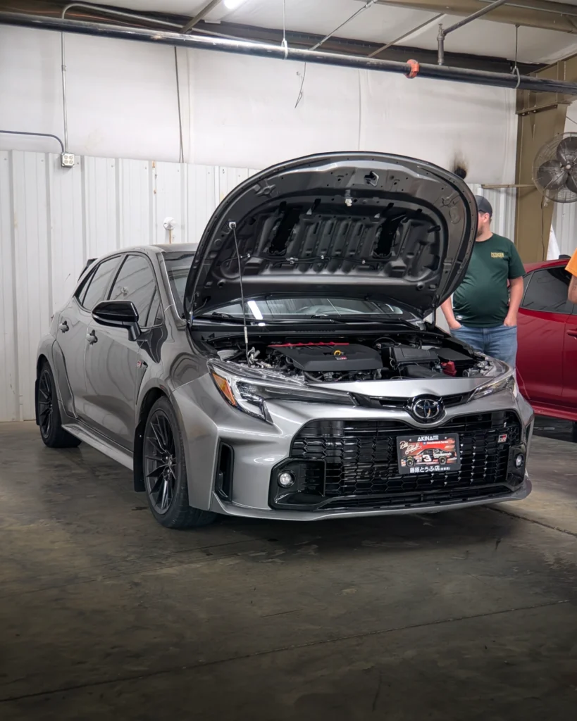Silver Toyota Corolla GR hatchback with hood open inside a garage space and two men standing nearby.