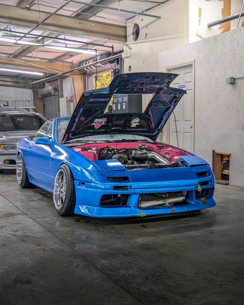 Blue Mazda RX-7 with hood open inside a garage, engine and pink engine bay visible
