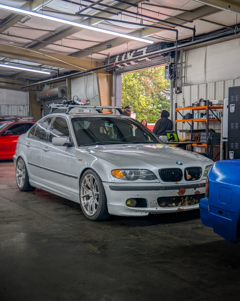 Silver BMW sedan parked indoors near garage door with people in background and a blue vehicle partially visible