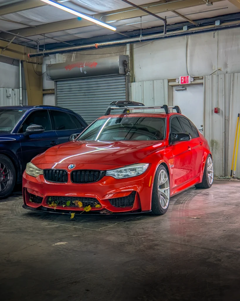 Red BMW sedan parked indoors with roof rack, alongside a blue SUV, front 3/4 view.