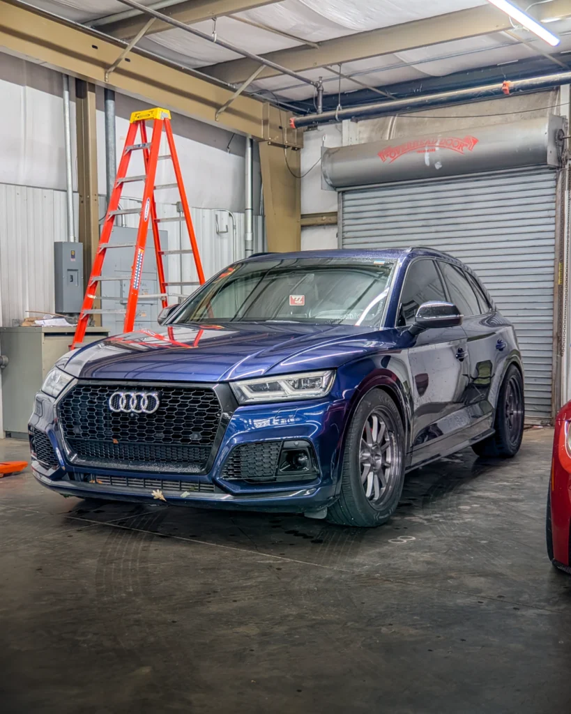 A blue Audi Q5 is parked inside a garage beside a red ladder and a closed roll-up door.