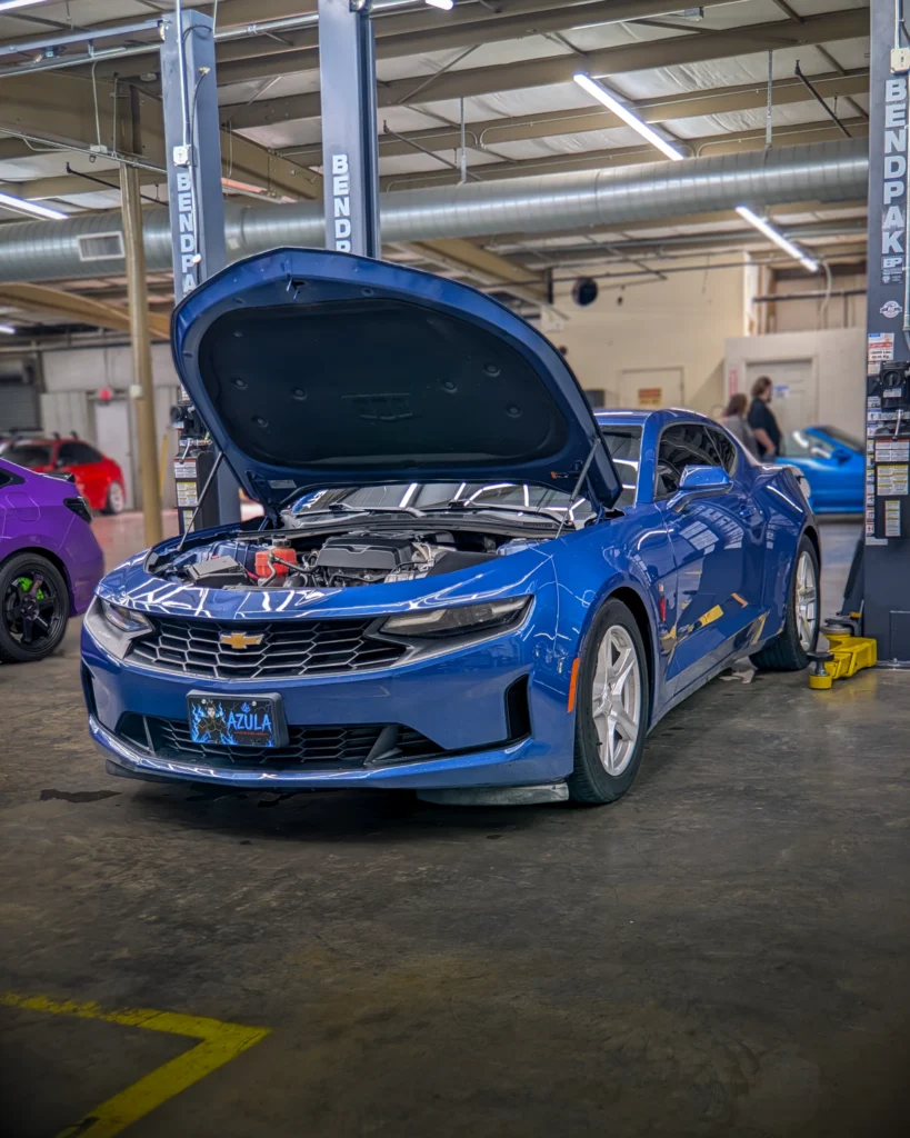 Blue Chevrolet Camaro with open hood in indoor garage with other vehicles and people in background