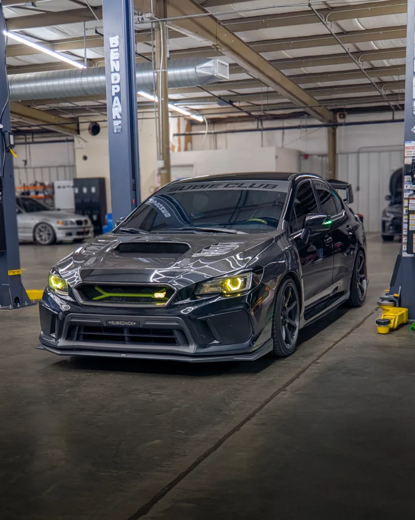 Black Subaru WRX STI with yellow headlights and carbon fiber hood in a garage workshop