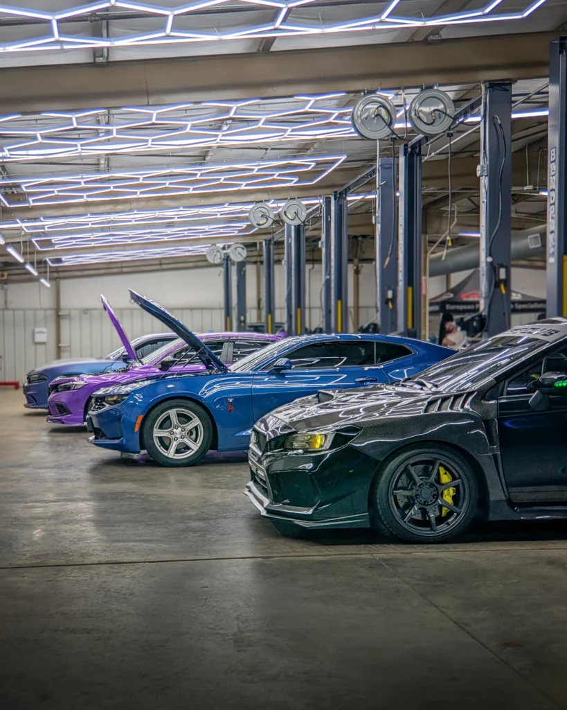 Three cars with open hoods in an indoor automotive workshop with overhead lighting and equipment lifts