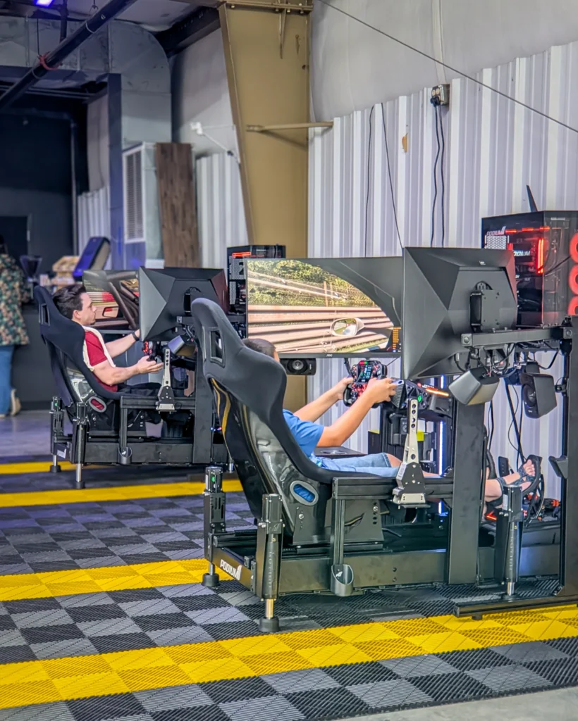 Two people seated at racing simulator rigs with steering wheels and large monitors inside a building with industrial decor.