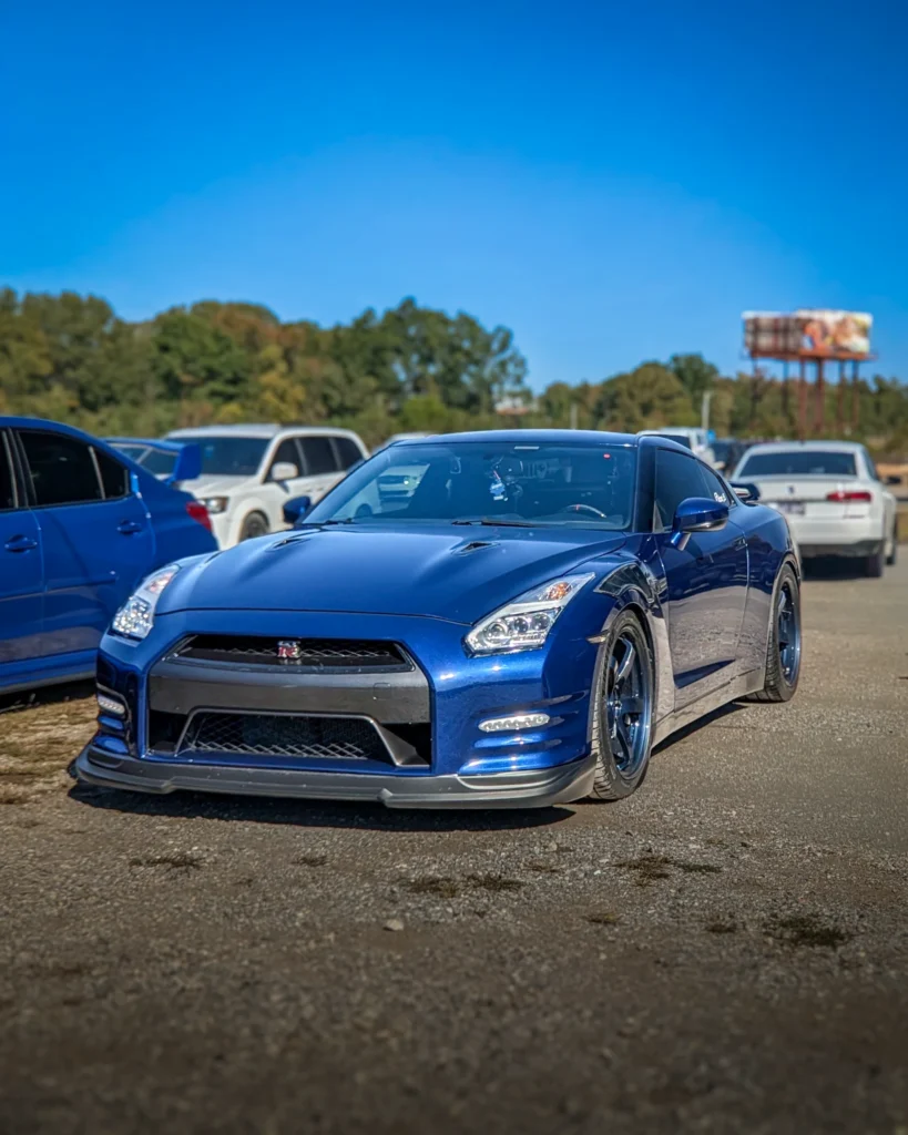 Blue Nissan GT-R parked in a lot with several other vehicles visible in the background under a clear blue sky