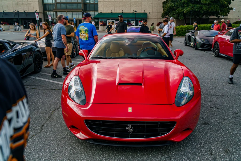 Red Ferrari California convertible front view with people and other cars in a parking lot at a car event