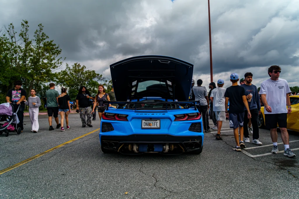 Blue Chevrolet Corvette with rear hood open parked outdoors among people under cloudy sky