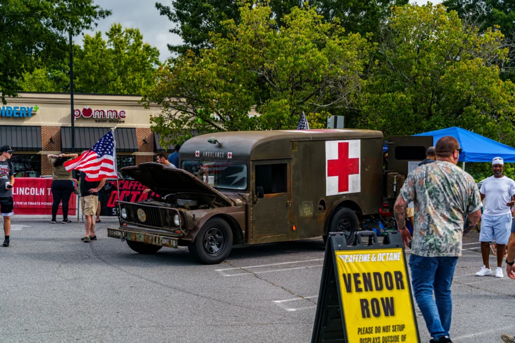 Olive drab military ambulance with red cross signs and hood open in a parking lot at a car event with people around