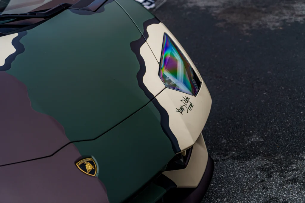 Close-up of front left headlight and hood of a camo-pattern Lamborghini Aventador with visible badge on asphalt.