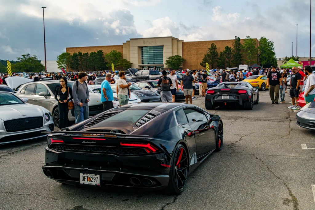 Black Lamborghini and white Bentley at a crowded car meet outside a shopping mall on a cloudy day