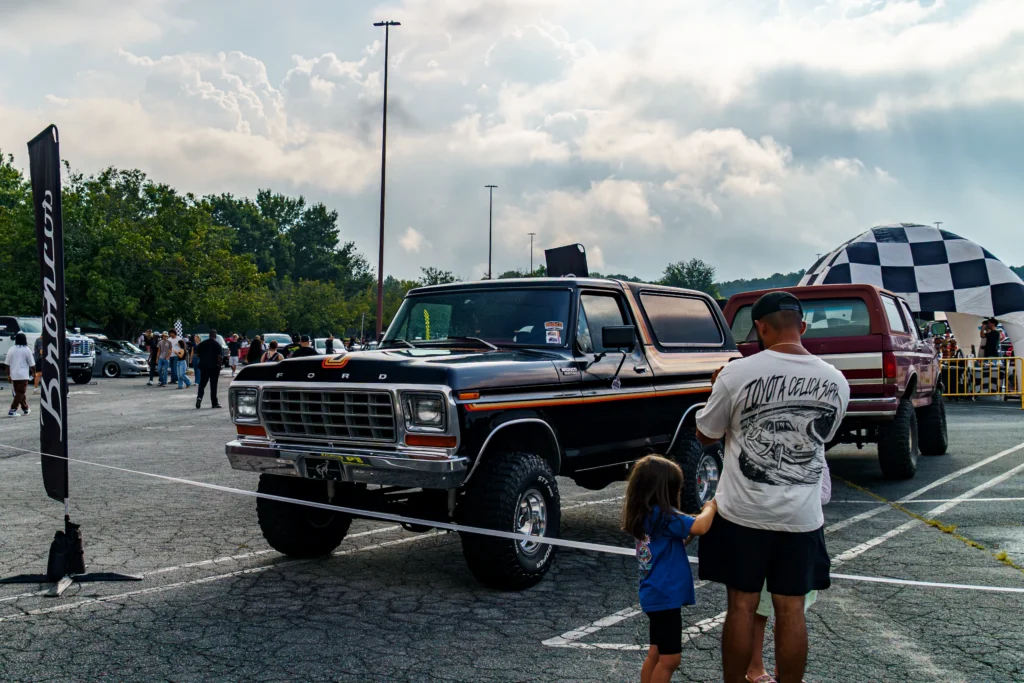 Black Ford Bronco with lifted suspension parked at an outdoor car event with attendees and a checkered inflatable arch in
