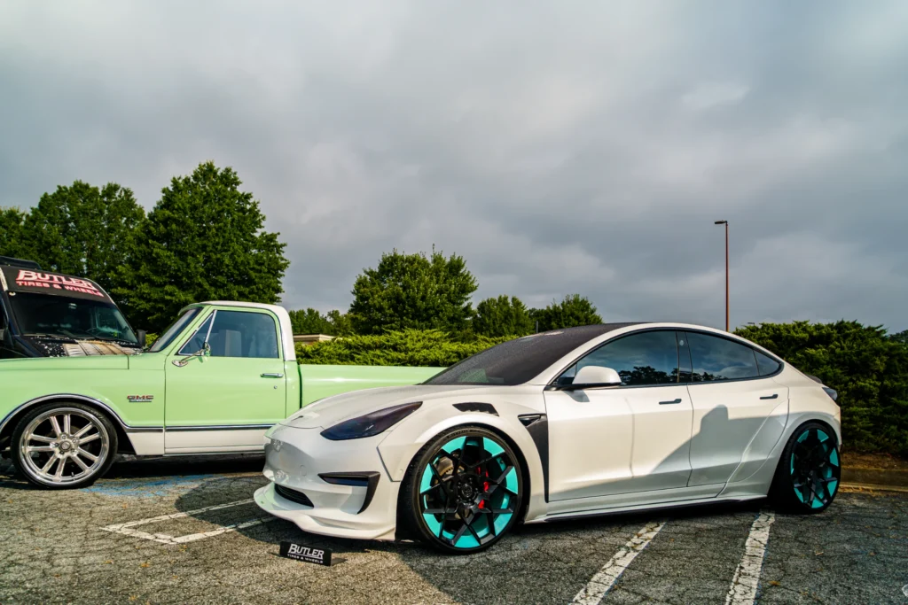 White Tesla Model 3 with black and turquoise wheels parked next to a light green GMC pickup truck in an outdoor lot with