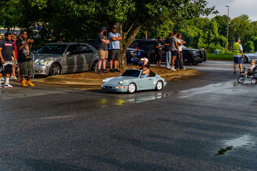 Small light blue Porsche 911 ride-on car driving on wet pavement with helmeted driver, crowd and parked cars in background.