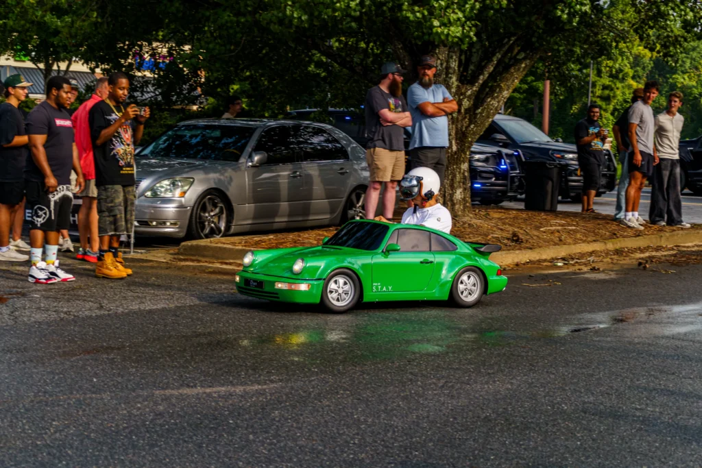 Small green Porsche 911 replica ride-on car with driver wearing white helmet and jacket on street near parked silver sedan