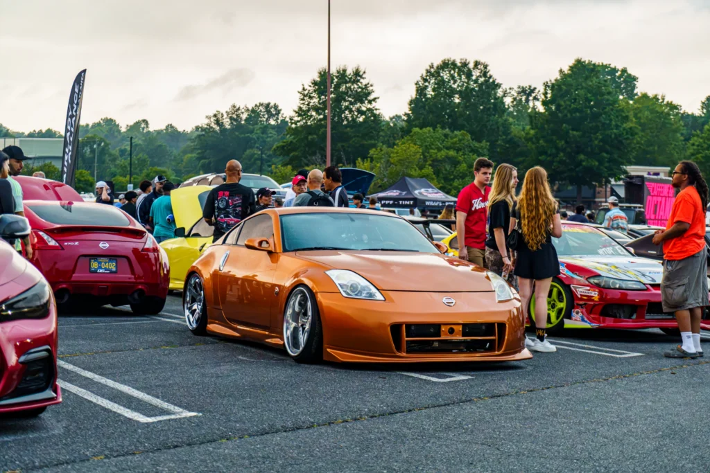 Orange Nissan 350Z parked among other cars at an outdoor car event with people walking around