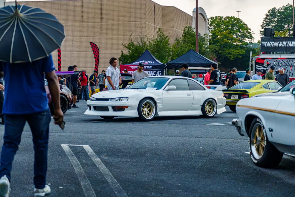 White Nissan 240SX coupe with gold wheels parked in a lot at a car meet with people and tents nearby.