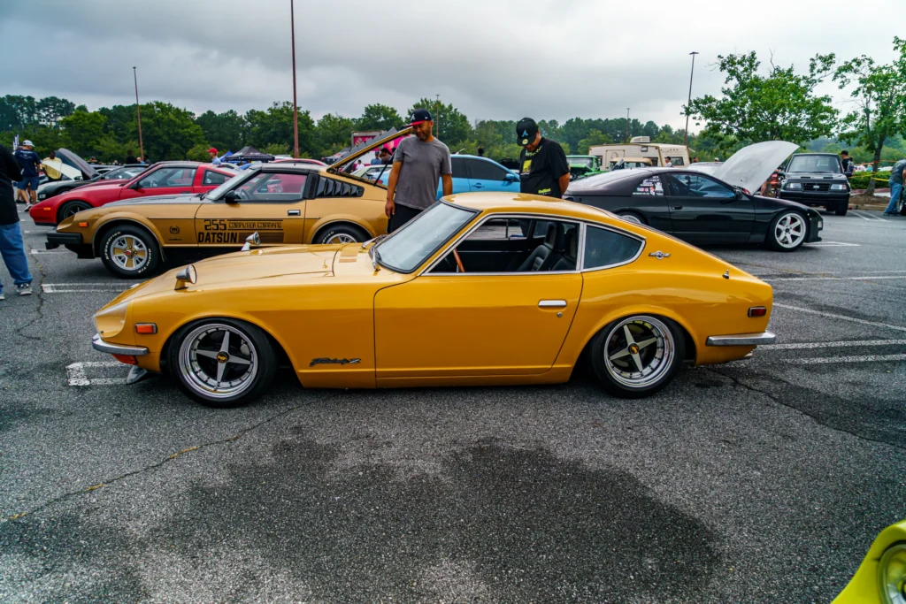 Yellow Datsun 240Z parked with other Datsun and Nissan cars at an outdoor car meet under cloudy sky.