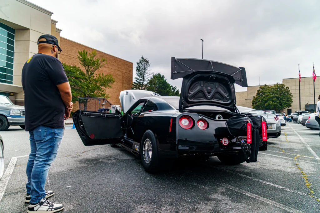 Black Nissan GT-R with open doors and rear hatch at a parking lot car event, with a man standing nearby and other vehicles