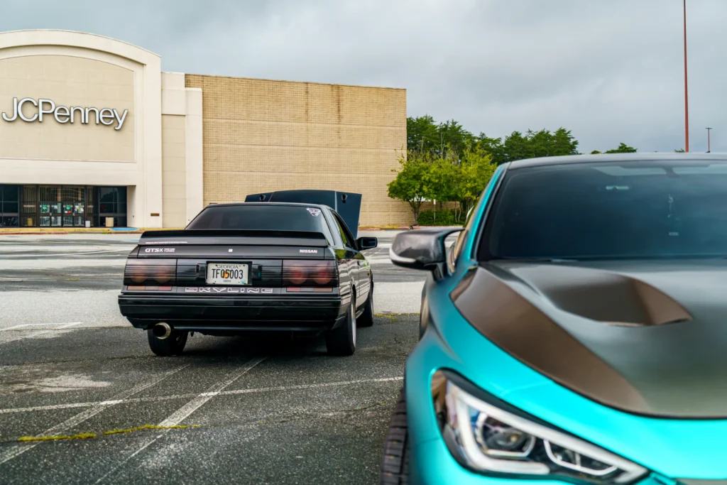 Black Nissan Skyline GTS-X with hood open parked in a lot near a teal car front corner and a JCPenney store