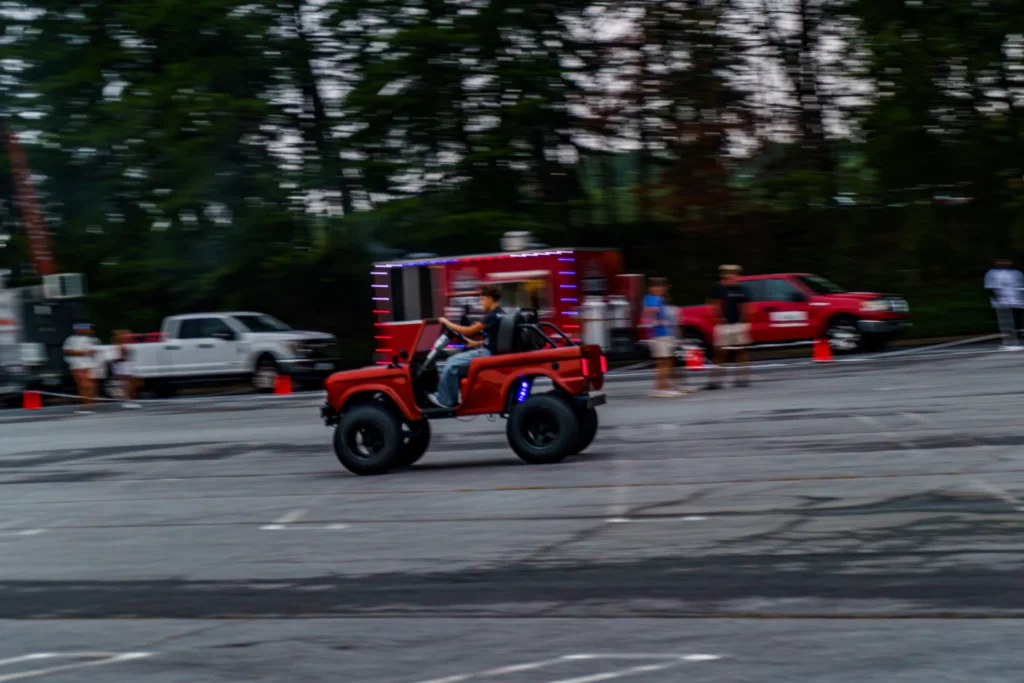 Red off-road vehicle with lifted suspension driving through a paved area with people and trucks in the background