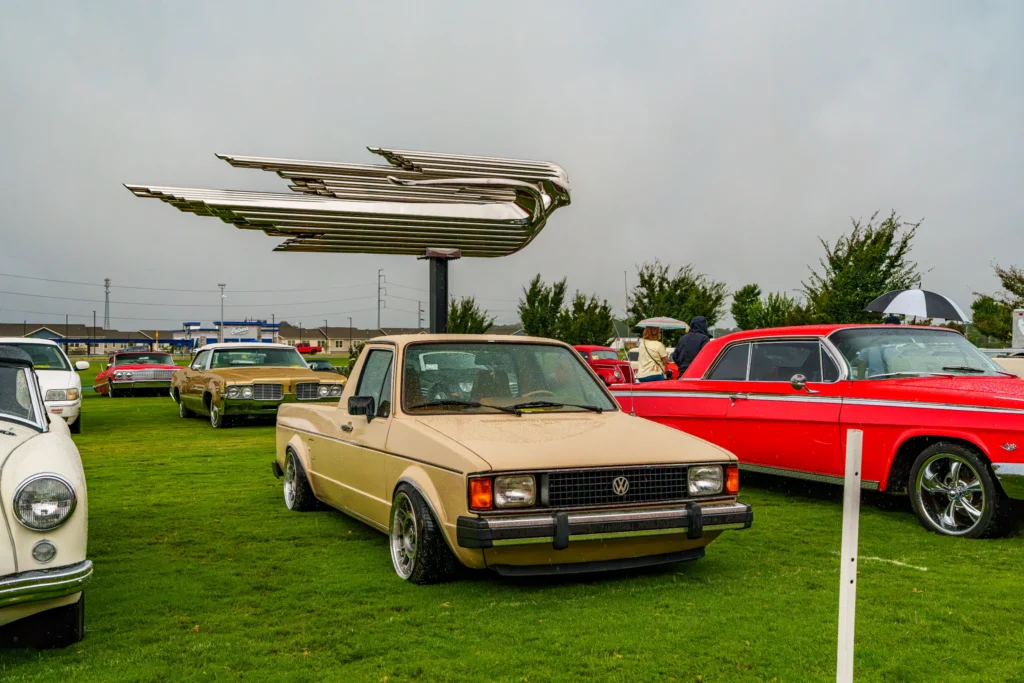 Beige Volkswagen Rabbit and red Chevrolet coupe parked on grass with rain and large chrome bird sculpture overhead