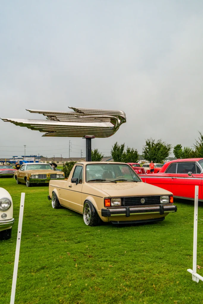 Tan Volkswagen Rabbit parked on grass at a car meet under large metallic bird sculpture with other classic cars in background