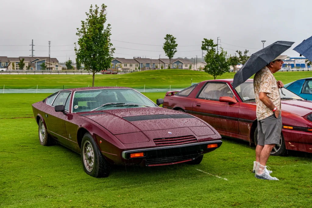 Maserati Khamsin parked on a grass field with a man holding an umbrella nearby on a rainy day