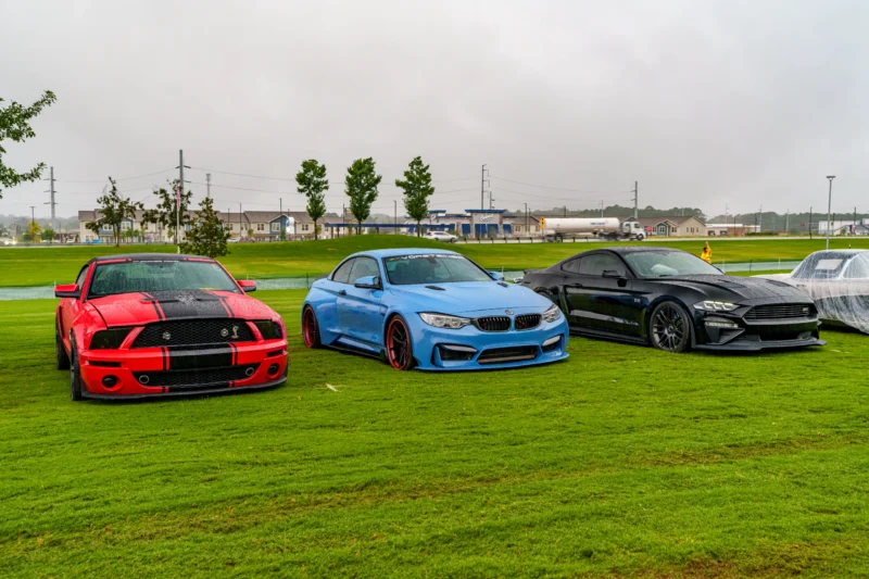 Red Ford Mustang Shelby GT500, blue BMW M4, and black Ford Mustang parked on grass in overcast weather at car event