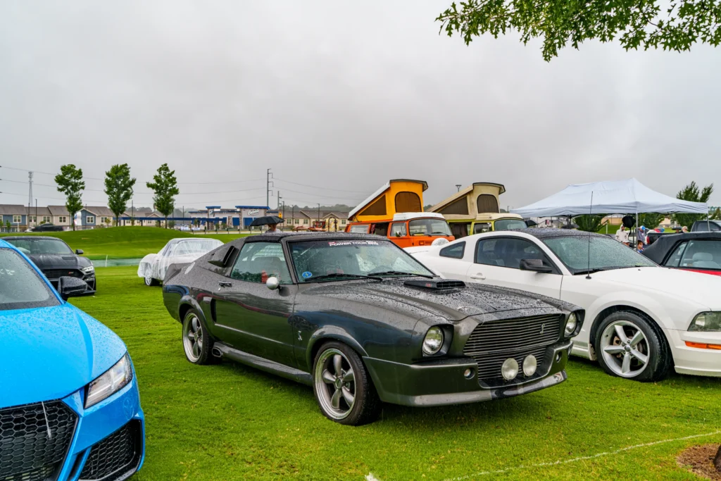 Black Ford Mustang II with a hood scoop and fog lights in a rainy car meet setting with other vehicles and tents nearby