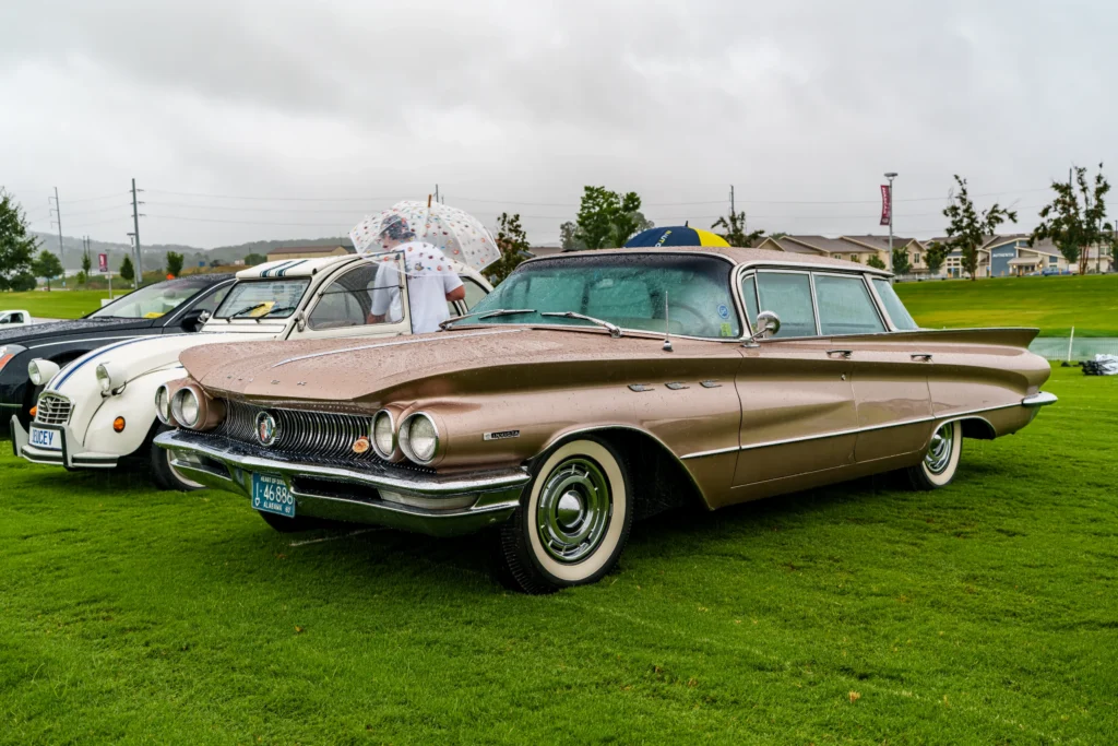 Beige Buick Invicta sedan parked on grass with raindrops visible, white car with blue stripes and umbrellas in background