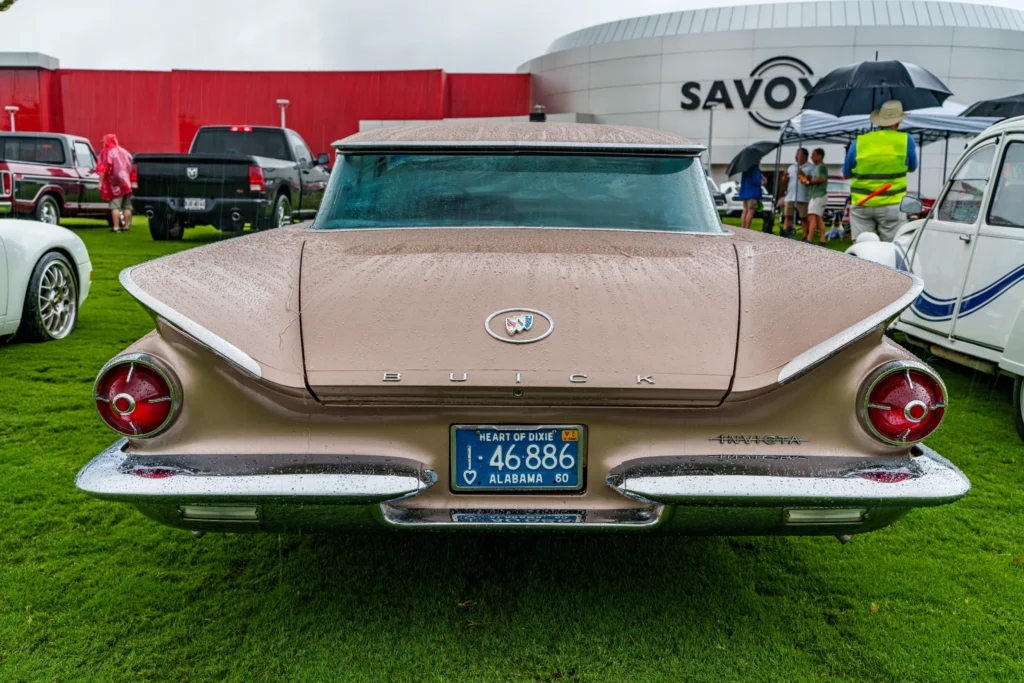 Rear view of a tan Buick Invicta parked on grass at a rainy car event with people and tents in the background.