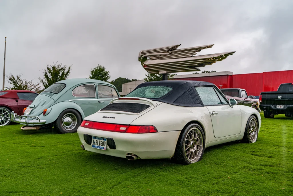White Porsche 911 Carrera convertible and light blue Volkswagen Beetle parked on grass with a large metallic sculpture and