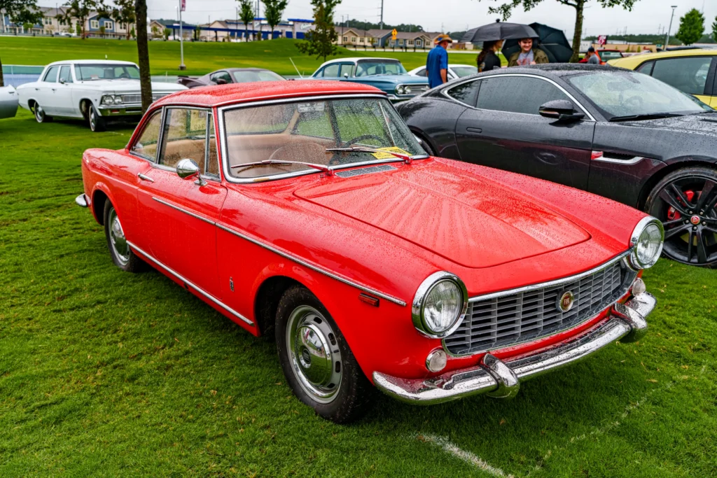 Red Fiat 1500 Coupe and parked cars on wet grass at an outdoor car event on a cloudy day