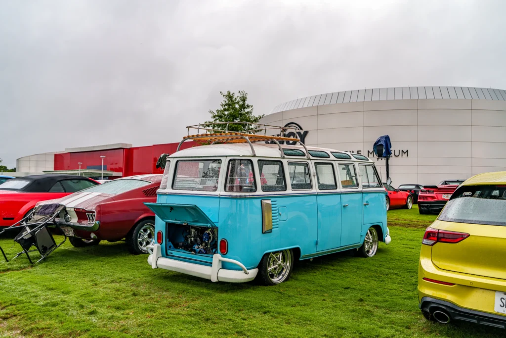 Light blue and white Volkswagen Type 2 van with rear engine open and parked on grass at a car event with other vehicles.