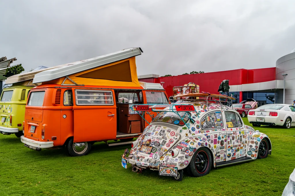 Orange Volkswagen Type 2 camper van with pop-up roof and heavily stickered white Volkswagen Beetle parked on grass at car