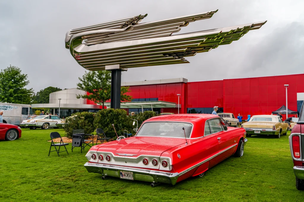 Red Chevrolet Impala parked on grass near a large chrome wing sculpture and other classic cars at an outdoor event