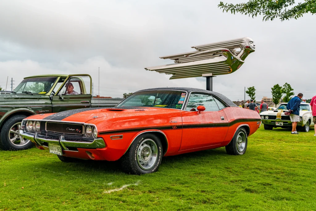 Orange Dodge Challenger R/T parked on grass with people and vintage truck nearby under steel sculpture