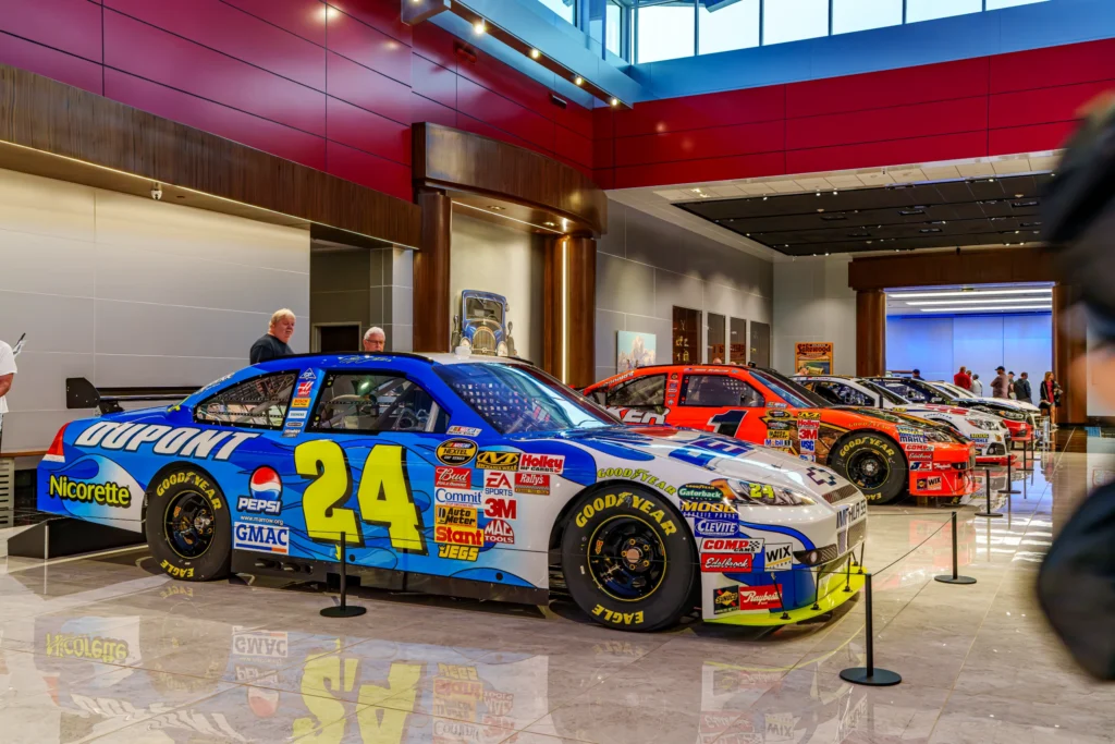 Four NASCAR stock cars parked indoors in a row with people near them and red and gray walls in the background