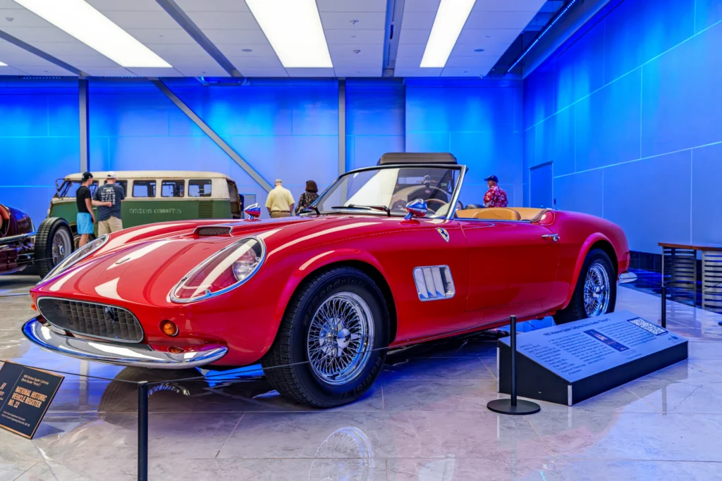 Red Ferrari 250 GT California Spyder convertible displayed indoors with blue-lit walls and visitors in background